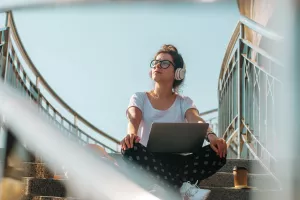 Female freelancer wearing headphones working on the laptop and drinking coffee. Sunny day in the city. Generation Z concept.<br>generacija Z, gen z, mladi, mladež, nove generacije, mladost, mlađi