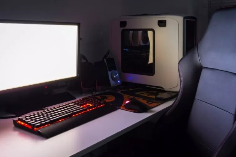 Custom built gaming computer with white screen, keyboard, mouse, joypad, gaming chair under low light. Selective focus.