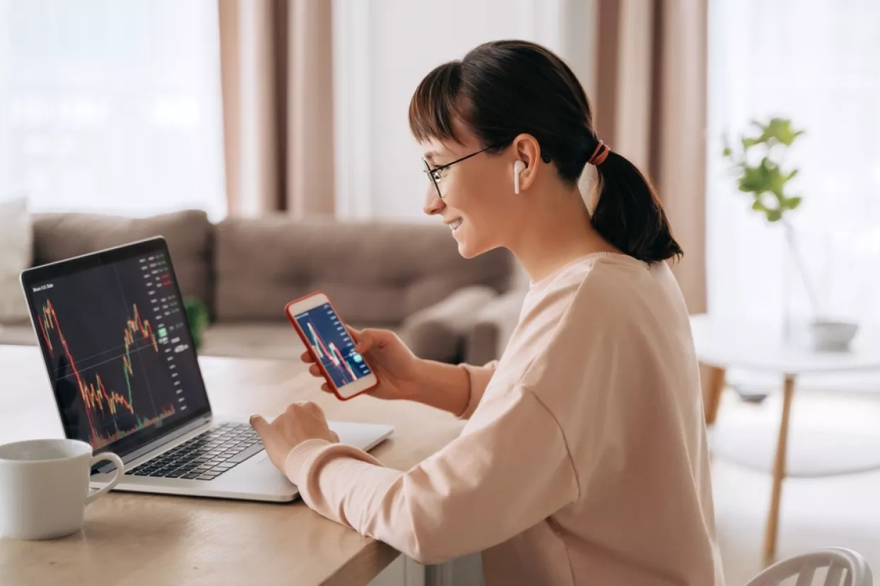 Smiling business woman trader analyst looking at laptop monitor, holding smartphone, wearing earphones. Investor broker analyzing indexes, trading online investment data on stock market graph at home<br>investiranje, mladi, studenti, ulaganje, učenje, burze