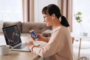 Smiling business woman trader analyst looking at laptop monitor, holding smartphone, wearing earphones. Investor broker analyzing indexes, trading online investment data on stock market graph at home<br>investiranje, mladi, studenti, ulaganje, učenje, burze