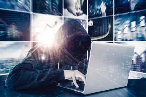 Double exposure of hooded hacker using a laptop while sitting with blurred virtual screen background<br>NFT prijevara, krađa