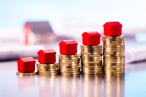 Small red houses standing on stacks of coins with blueprints and architectural model in the background.<br>najamnina, cijene, cijene stanova, cijena stana, stanovi, nekretnine, kuće, kuća, rast