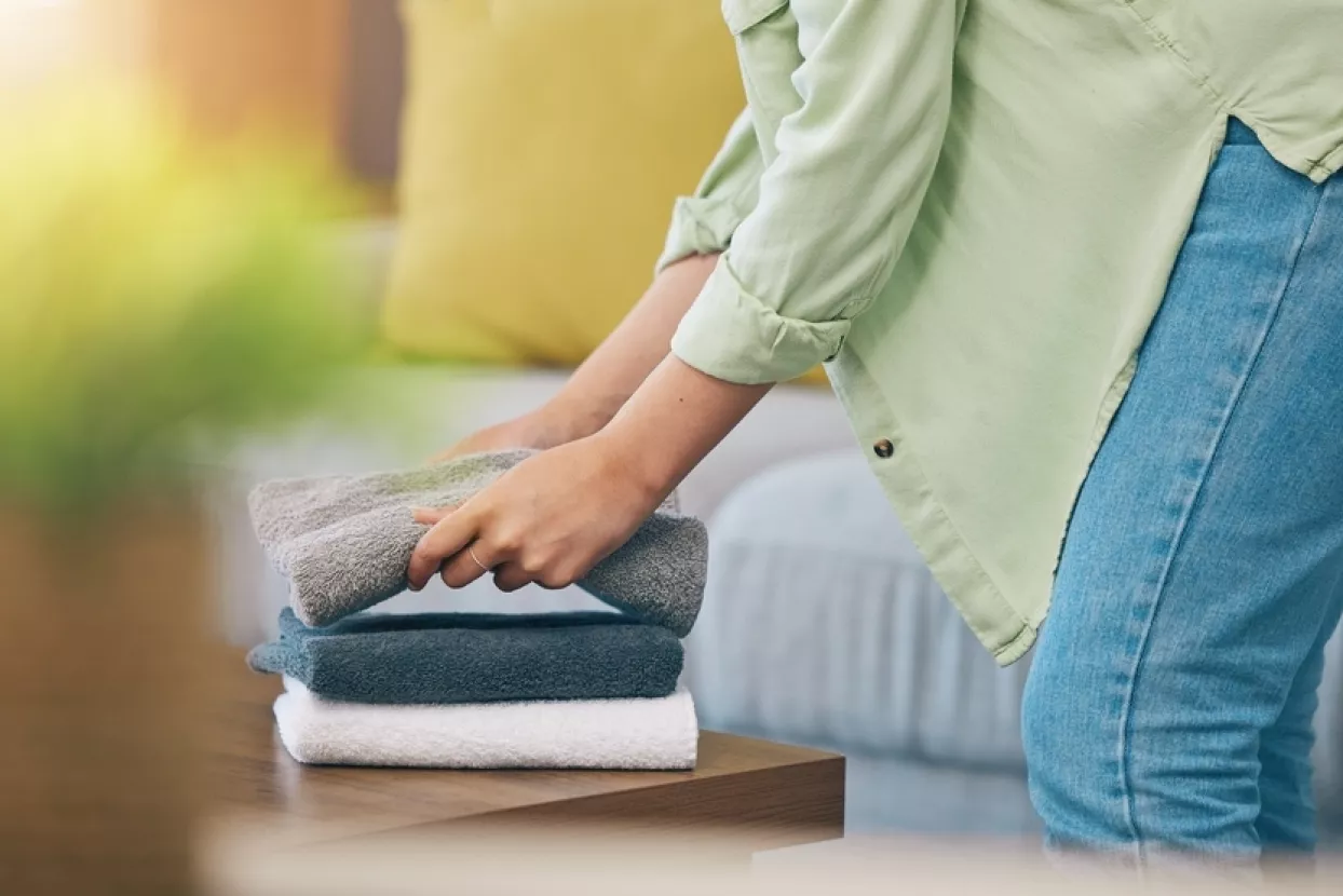 Hands, home and fold laundry on table for hygiene, cleaning and soft clothes for housework in living room. Woman, fabric and cotton towel on desk for organized house, service and routine in apartment
